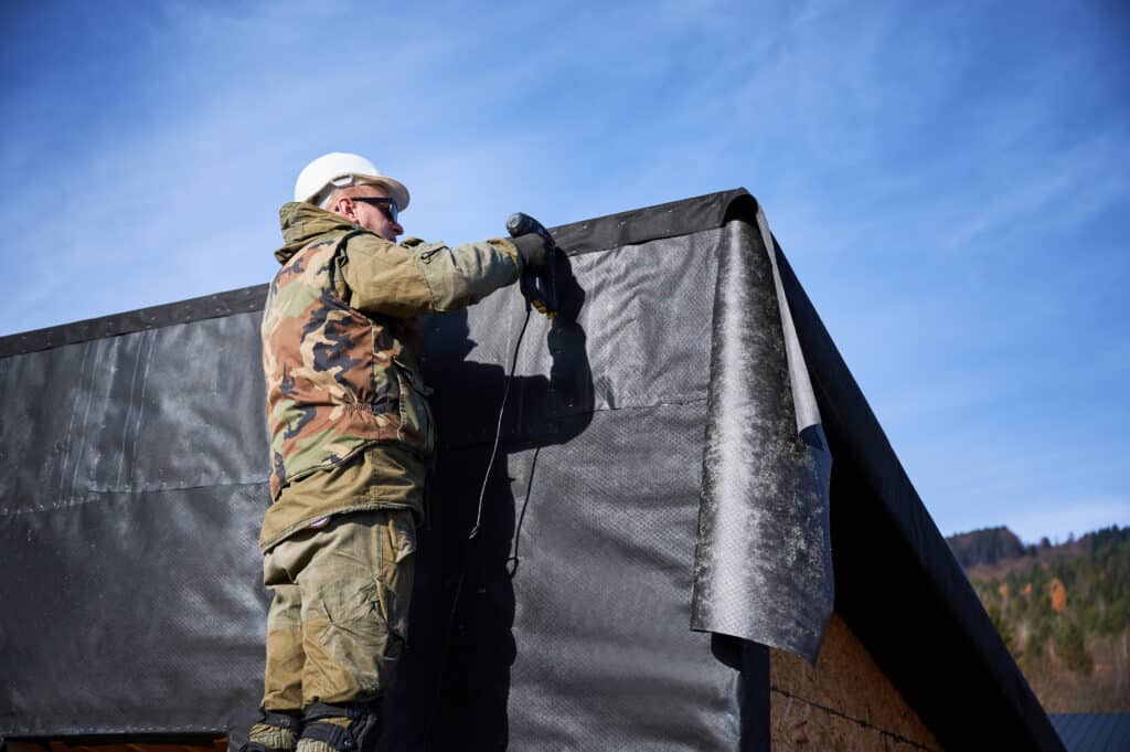 Male builder installing waterproof membrane on the wall of future cottage. Man worker building wooden frame house with blue sky on background. Carpentry and construction concept.